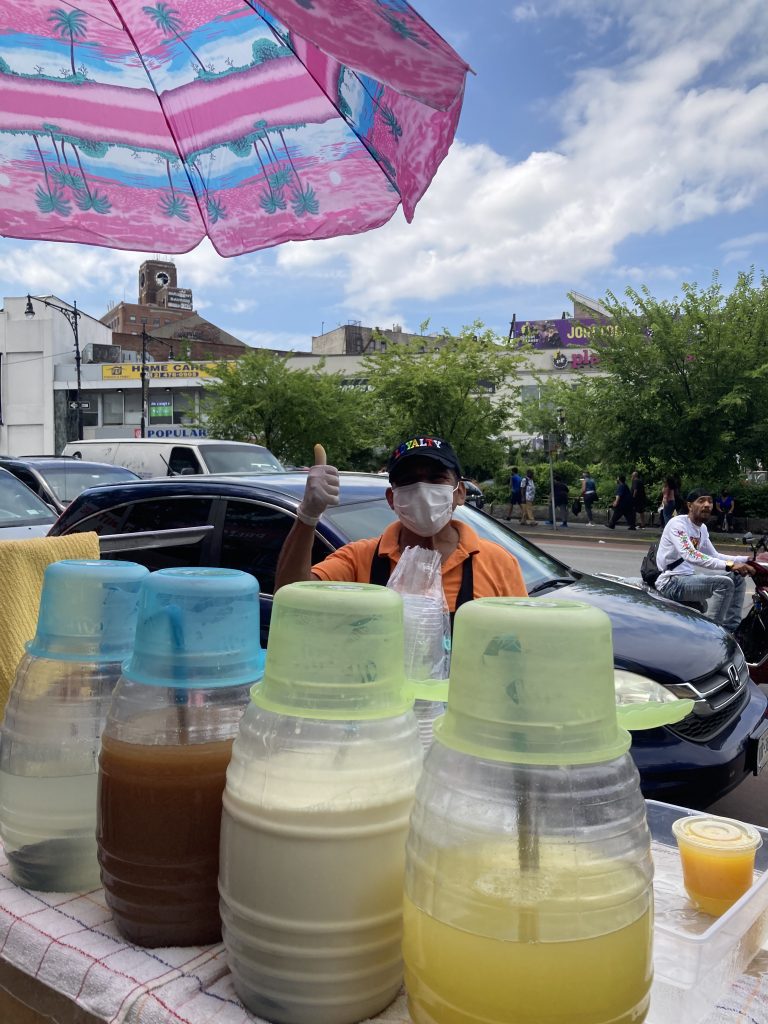 Lucio stands on a street in the Bronx wearing a mask with the drinks he sells in front of him.